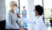 Image of African American doctor consoling her mature patient in waiting room at medical clinic. Both of them are wearing protective face masks. Image of African American doctor consoling her mature patient in waiting room at medical clinic. Both of them are wearing protective face masks.